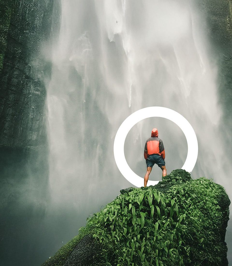 A person in a red jacket stands on a moss-covered rock, facing a towering waterfall, with a glowing white circle framing them.