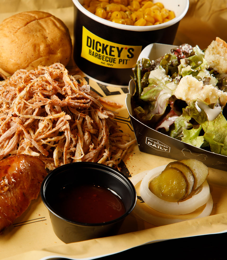 A close-up of a barbecue meal from Dickey's Barbecue Pit, featuring pulled pork, grilled chicken, salad, pickles, and a side of corn.