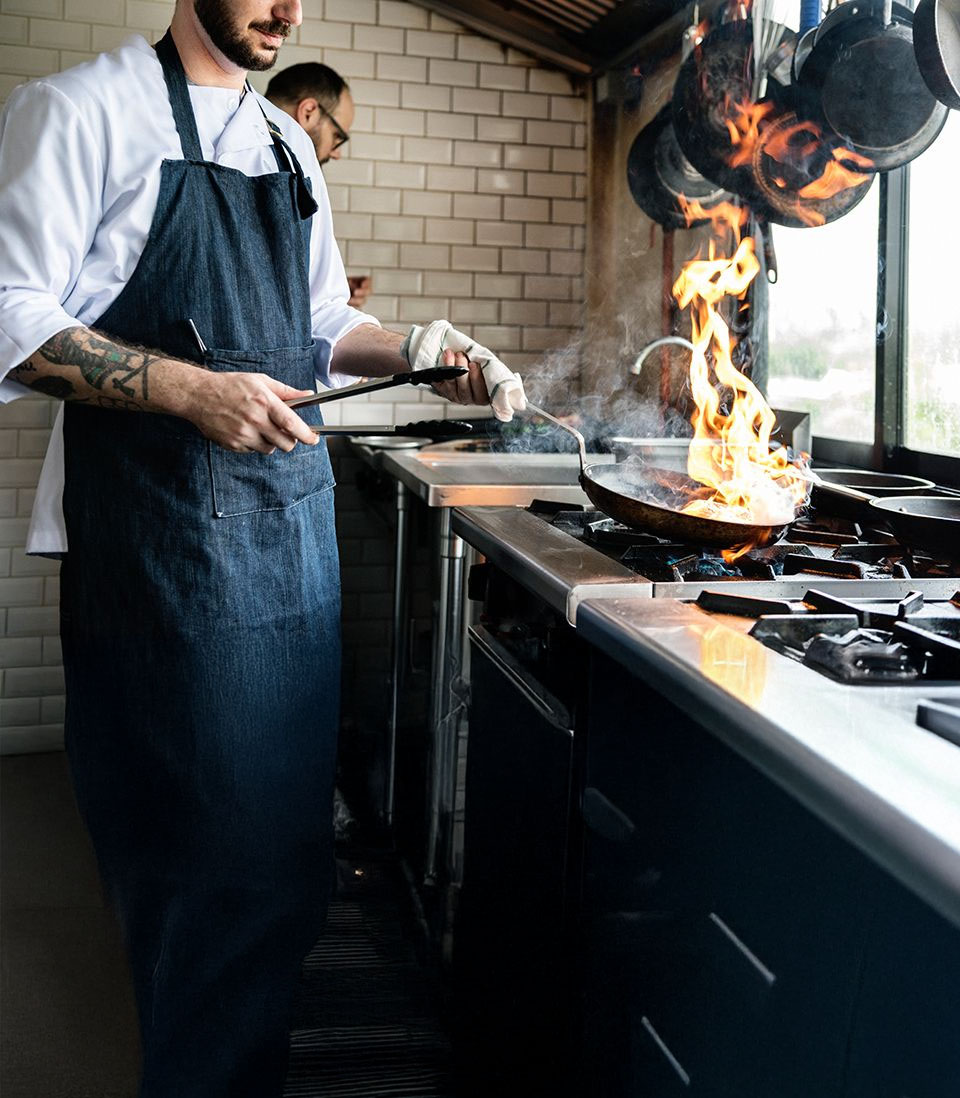 A chef in a professional kitchen, wearing a denim apron, skillfully handling a flaming pan over a gas stove, with another chef working in the background.