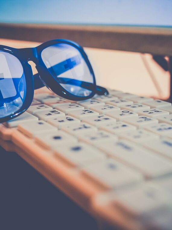 A pair of blue-tinted sunglasses resting on a white keyboard, reflecting the Facebook logo from a computer screen in the background.
