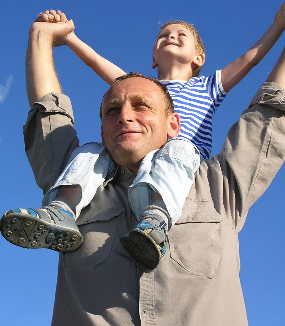 Father carrying smiling child on shoulders under blue sky.