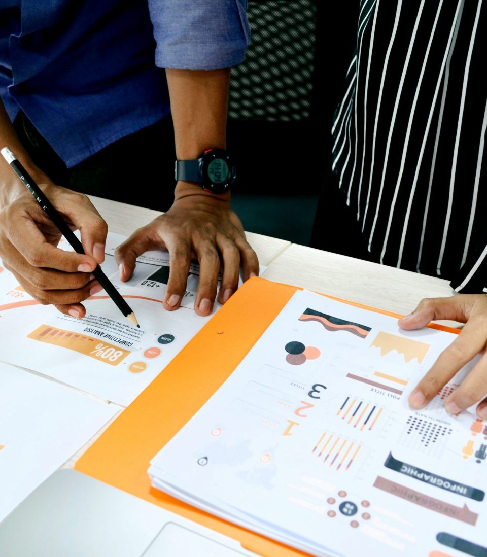 Document on a table displaying various data visualizations, with one person’s head leaning over the papers and another person’s hand pointing to a specific visual.