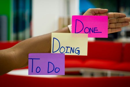 An outstretched hand in the foreground with three sticky notes on it: a purple note labeled "To Do," a yellow note labeled "Doing," and a pink note labeled "Done." The background shows an office setting.