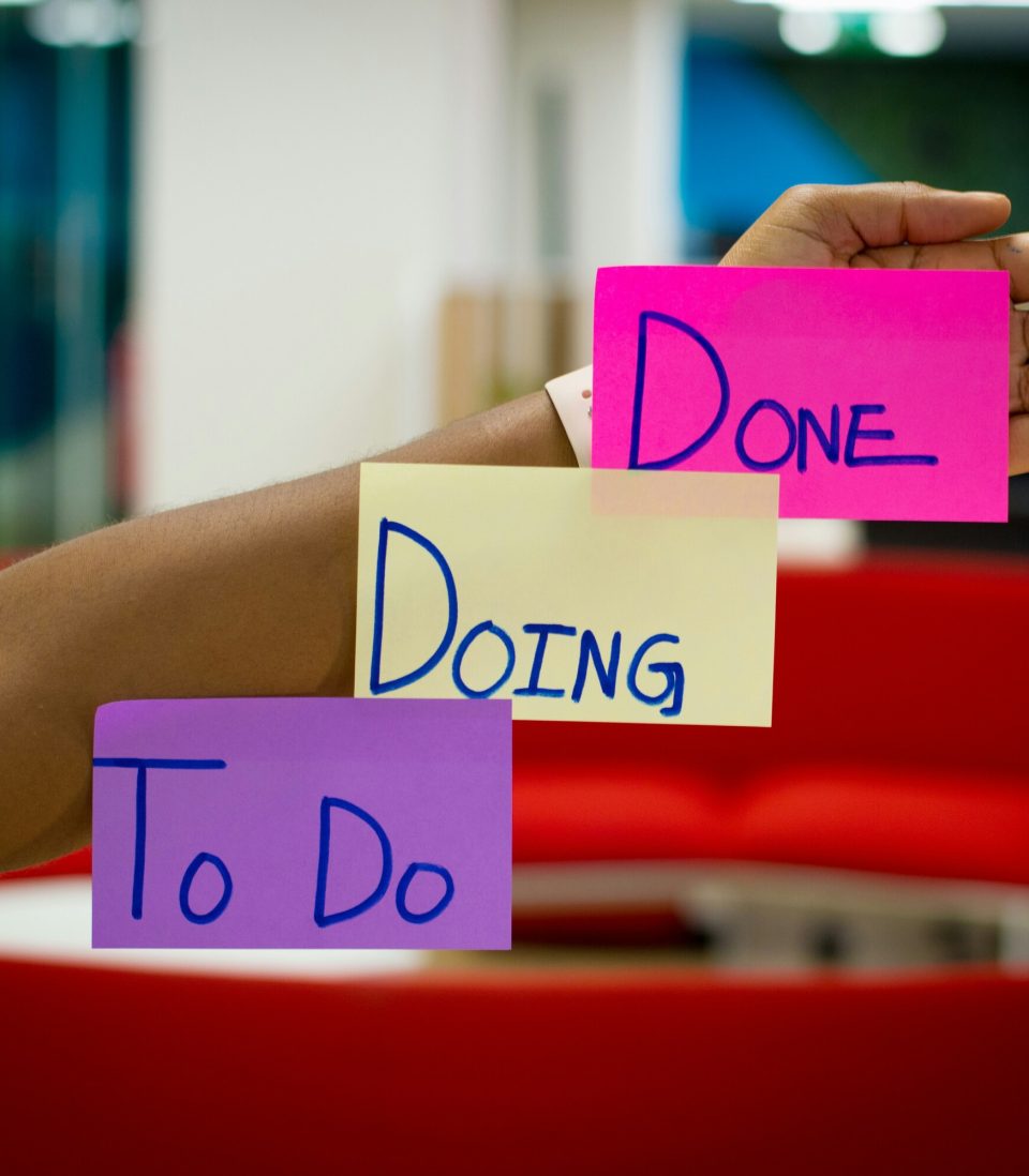 An outstretched hand in the foreground with three sticky notes on it: a purple note labeled "To Do," a yellow note labeled "Doing," and a pink note labeled "Done." The background shows an office setting.