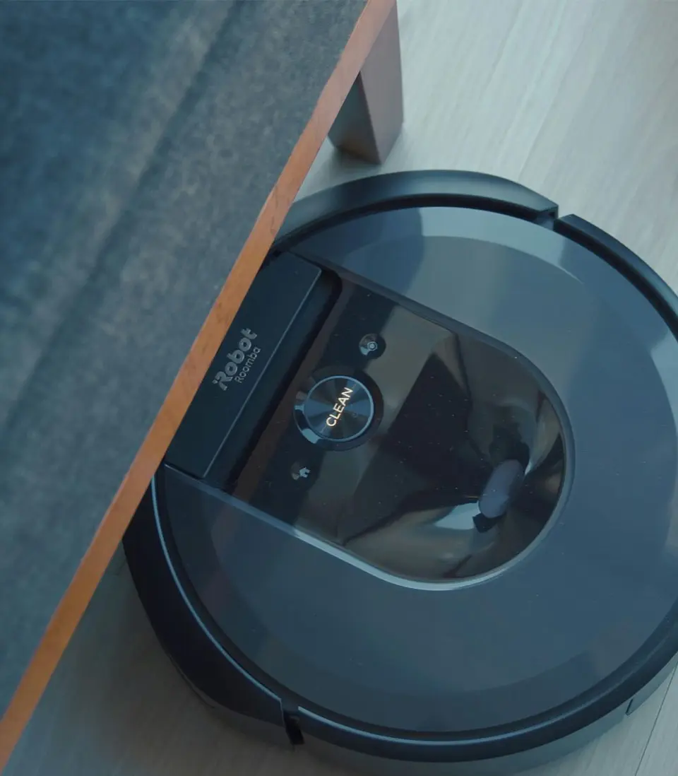 A robotic vacuum cleaner operating under a piece of furniture on a light wooden floor.