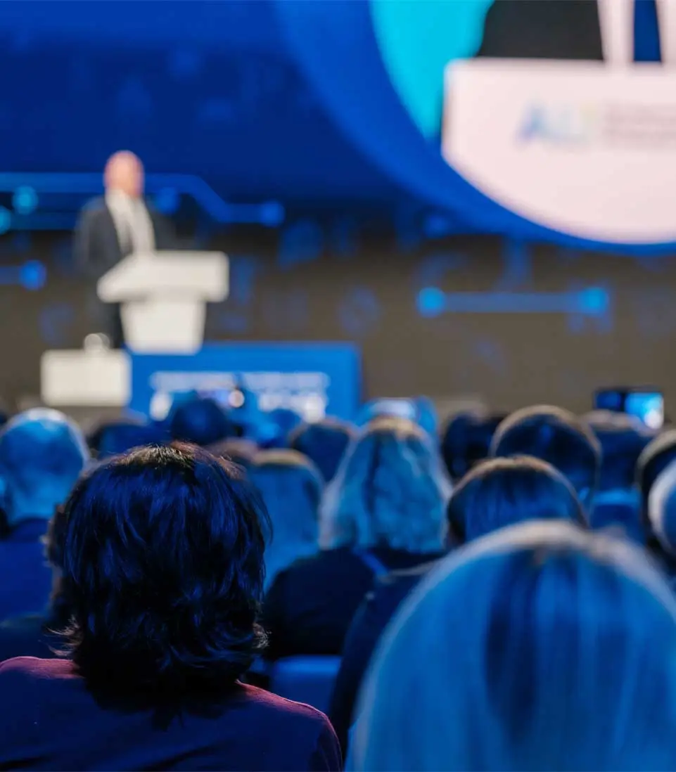 Audience seated at a conference watching a speaker on stage.
