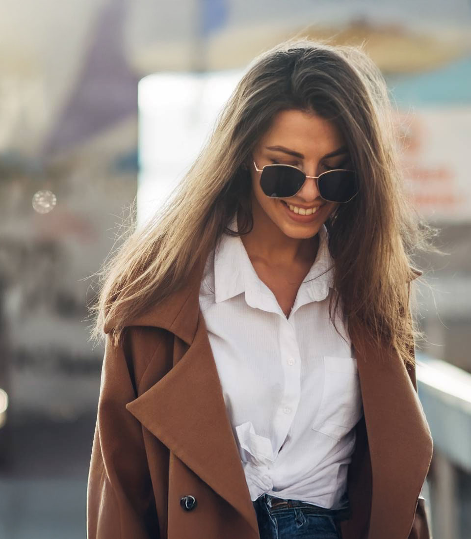 A stylish woman wearing sunglasses and a brown coat smiles while walking outdoors.