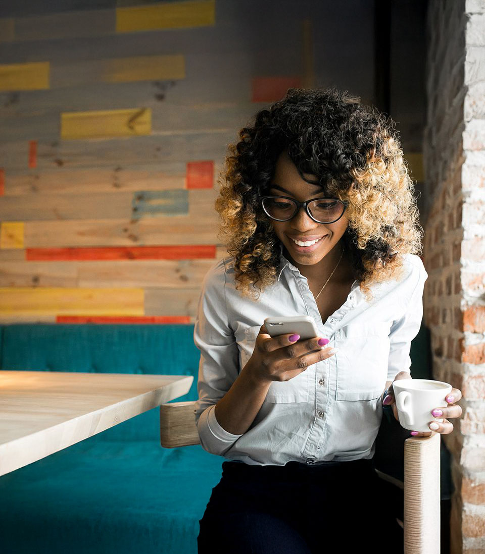 A woman in a café, smiling while looking at her phone and holding a coffee cup.