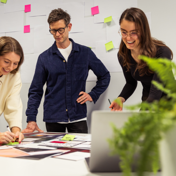 Alt text: Three colleagues collaborate in a creative workspace, smiling and engaged over design materials. A whiteboard with sticky notes is in the background, and a green plant is in the foreground.