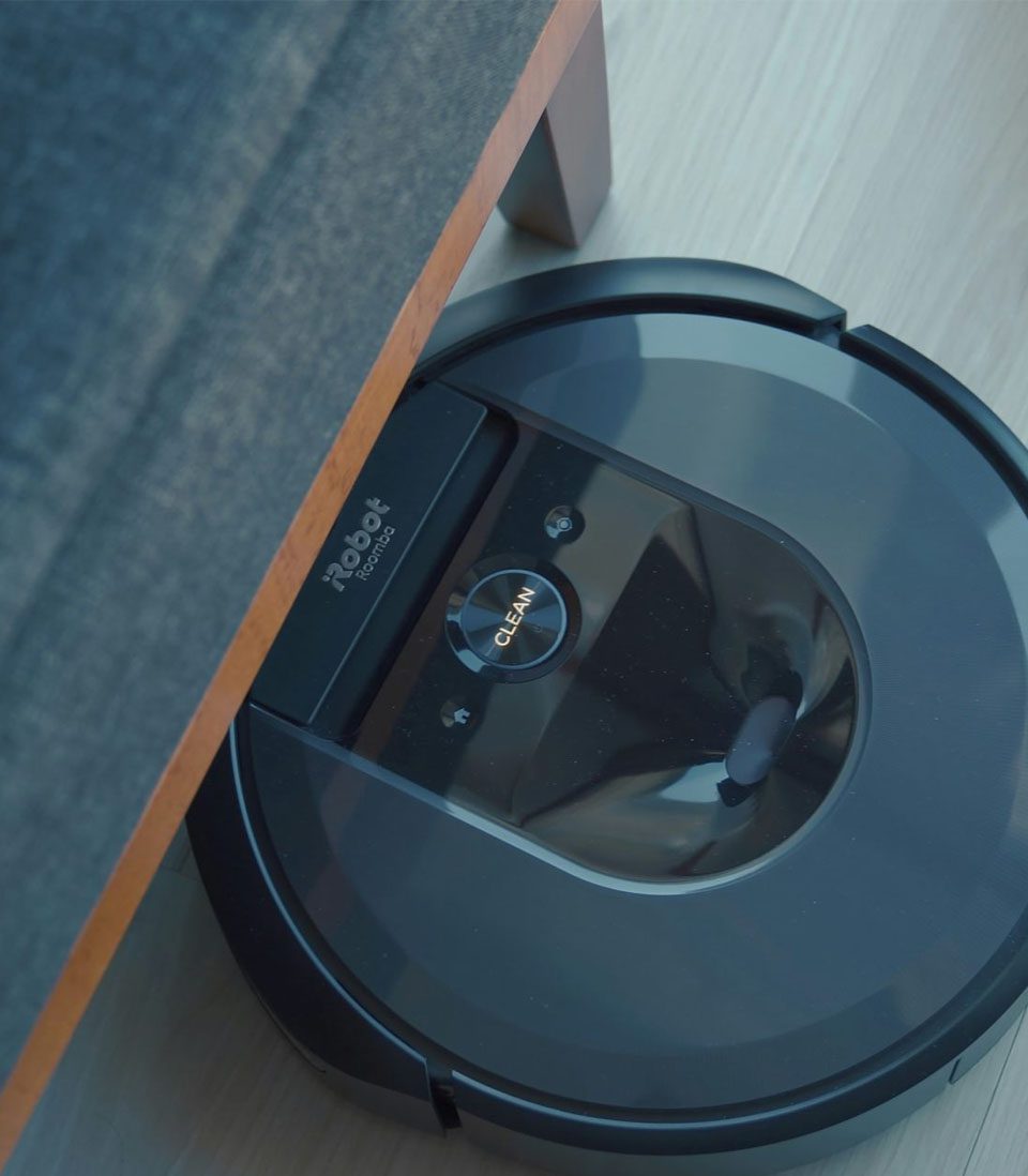 A robotic vacuum cleaner operating under a piece of furniture on a light wooden floor.