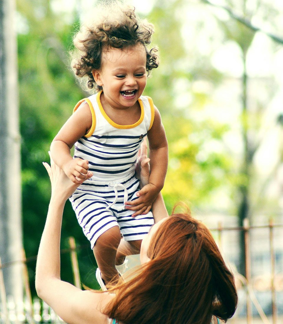 A joyful young child with curly hair is lifted into the air by a woman outdoors, both smiling.