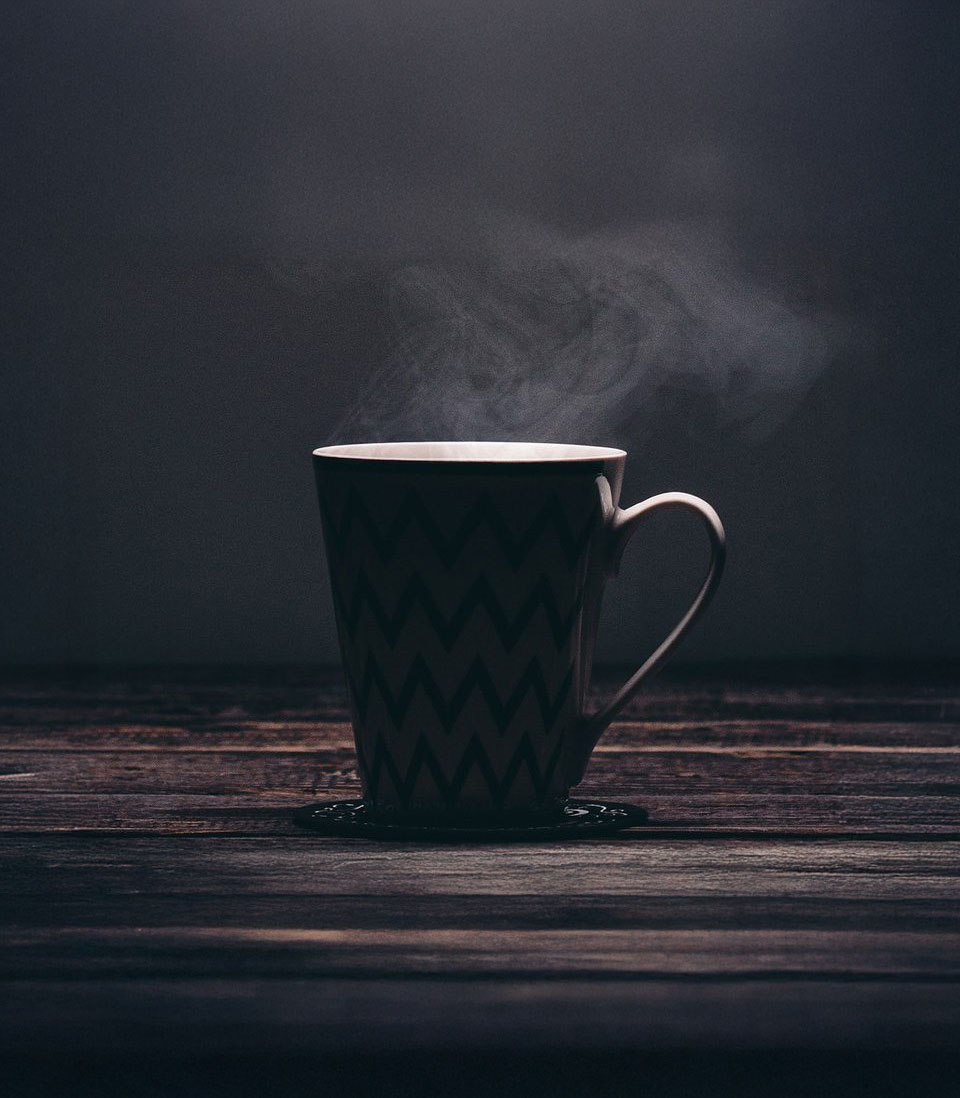 A steaming cup of coffee sits on a dark wooden table, with moody lighting and a minimalist background.