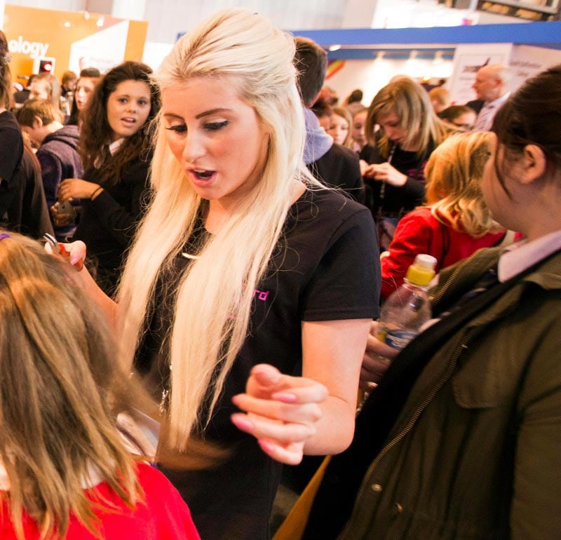 A hairstylist interacts with a young attendee at a lively event, showcasing techniques in a hands-on demonstration.