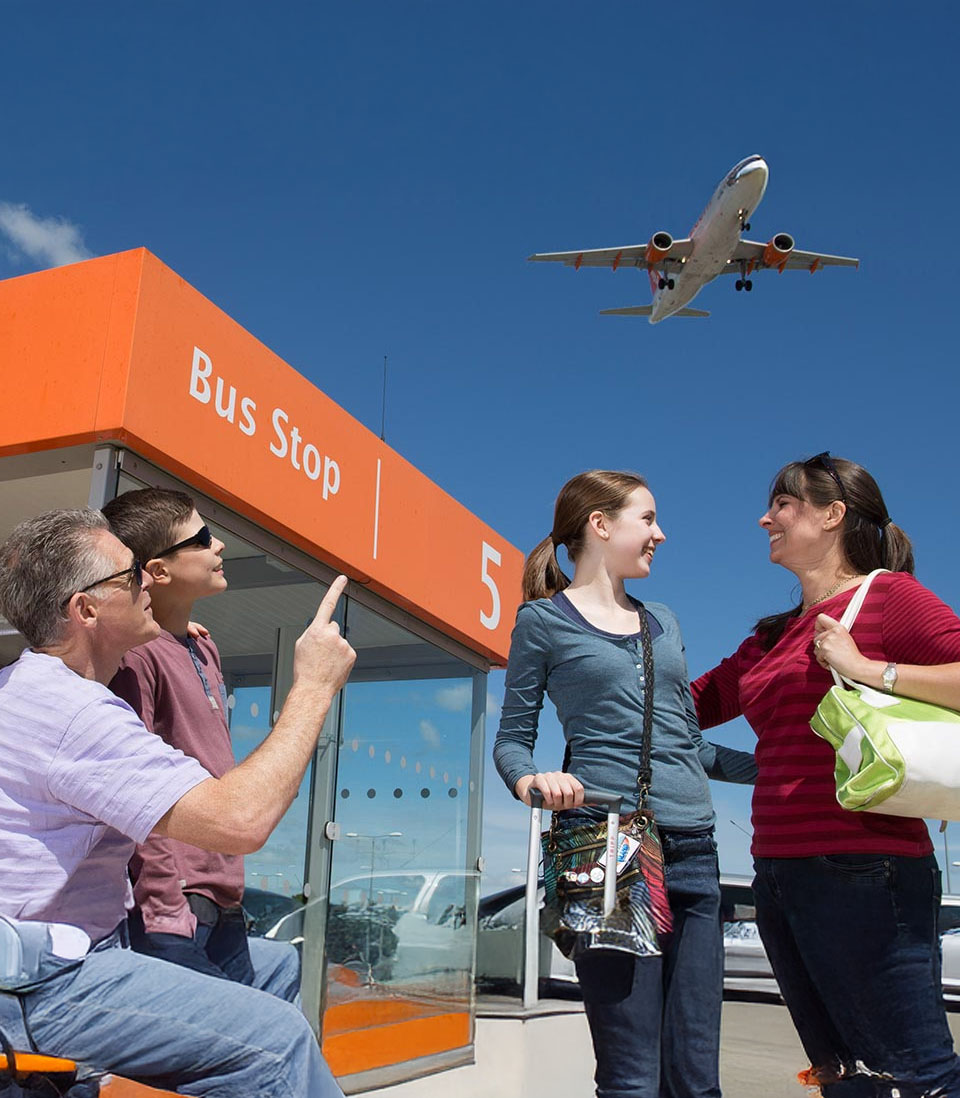A family waiting at an airport bus stop, with a man pointing at a plane flying overhead, while a mother and daughter smile at each other.