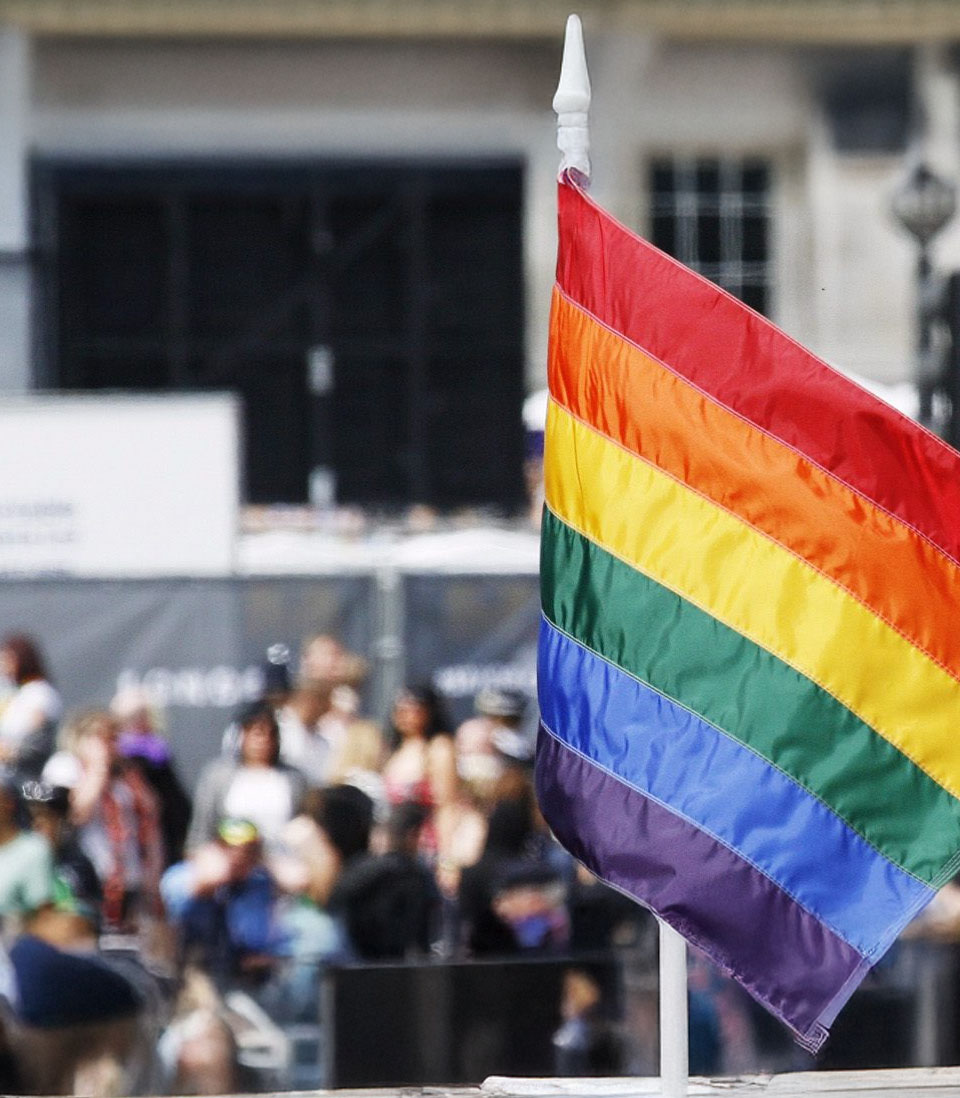 A rainbow Pride flag waving in the foreground with a crowd gathered in the background.