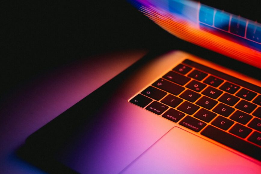 Half-closed laptop on a table in the dark with colorful light reflecting from the screen on the keyboard.